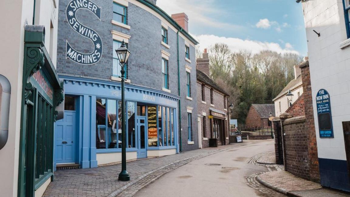 A curved street with Victorian buildings on both sides. one with a brightly painted blue front and the words "Singer Sewing Machines" on the wall.