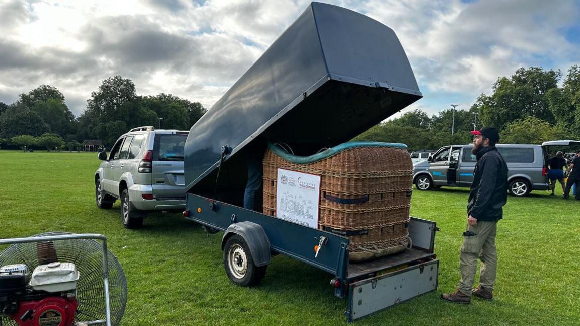 A balloon basket in a trailer hitched to the back of a 4x4. A bearded man in khaki trousers, dark jacket and black cap with a red peak is stood behind and to the right of it