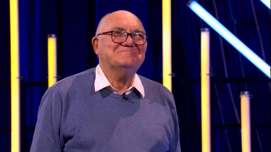 Harry Bowden looking off camera. He is smiling and behind him is a black and blue background with coloured neon lights. He has a bald head and is wearing a white shirt and blue jumper.
