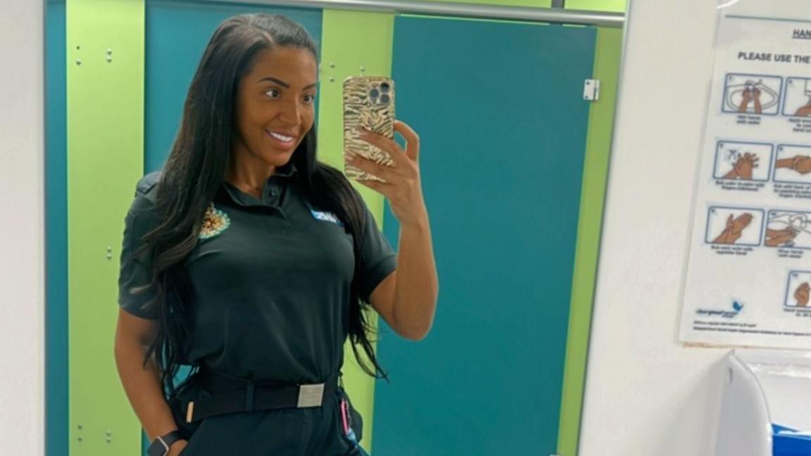 A girl taking a picture in a bathroom mirror. She is wearing a forest green paramedics uniform which is a short-sleeve polo top and long trousers with a black belt. She has long black hair is smiling into the camera. 