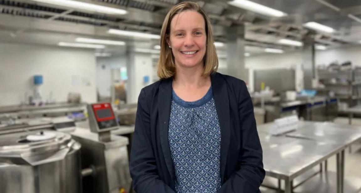 Susie Richardson, standing in a prison kitchen, smiling at the camera. She has shoulder-length blonde hair and is wearing a blue blazer with a blue and white top