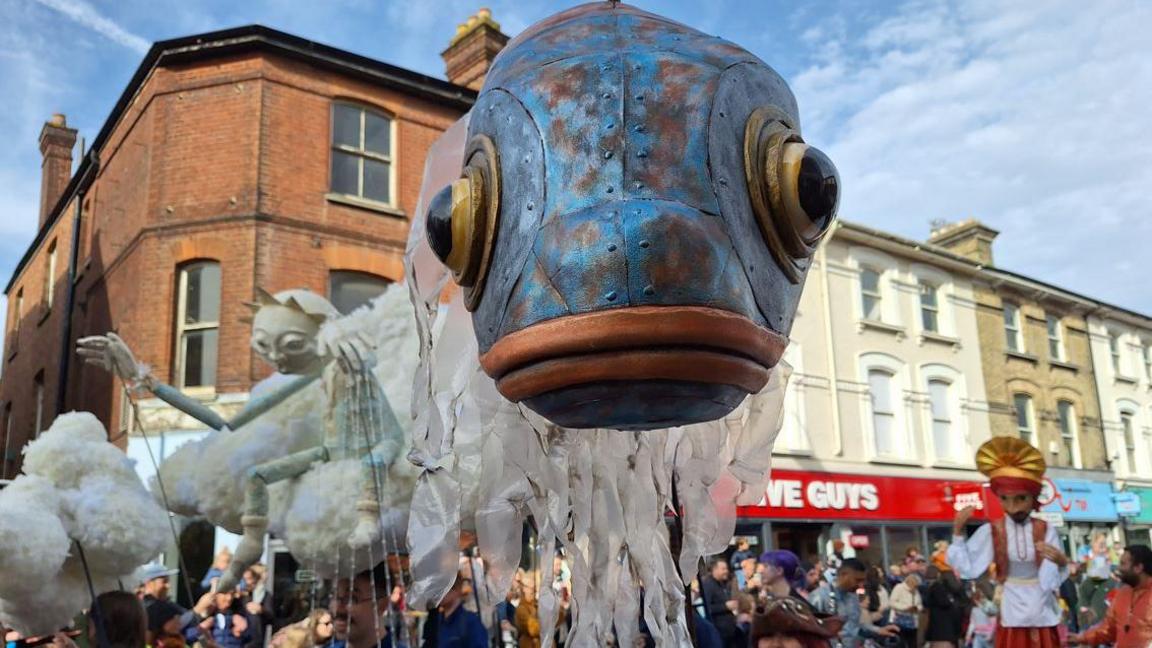 A giant fish puppet parading along Tunbridge Wells street with other puppets in the background