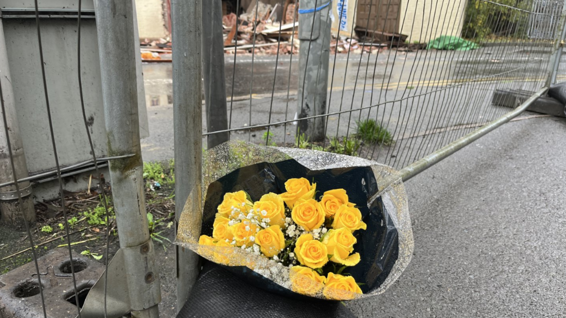 A bouquet of yellow roses is laid by a metal fence, with rubble and the foot of the house pictured in the background.