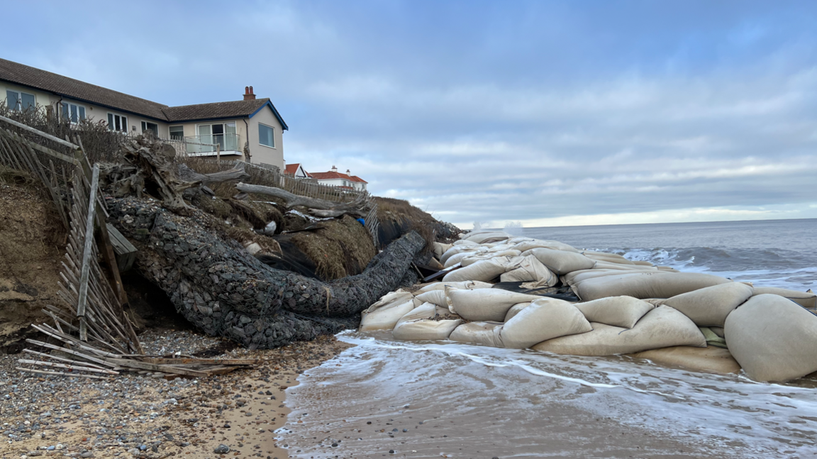 A home on the edge of an eroding cliff in Thorpeness. There is a big cluster of geobag sea defences on the beach in front of it. The tide is washing in.