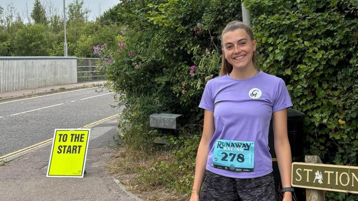 A woman in a purple running top and black running shorts smiles at the camera by a hedge.