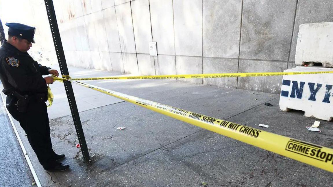 Police officer ties yellow caution tape around crime scene that includes bloodied side walk 
