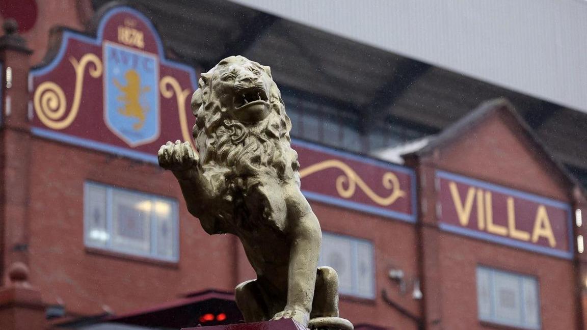 A statue of a lion with its paws reared at the front is in front of Villa Park, a red brick wall with AVFC painted on it.