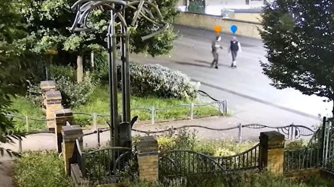 A CCTV image capturing two people talking in the street. The entrance to a park is nearby.