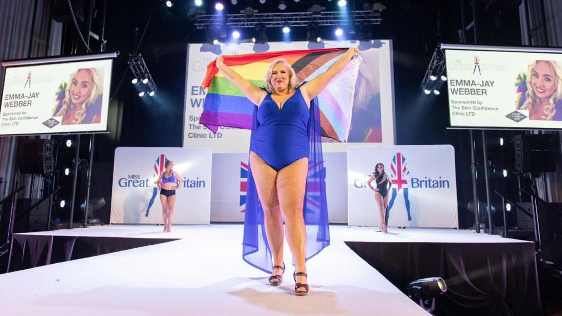 A woman wearing a blue bikini holding a rainbow pride flag on a catwalk