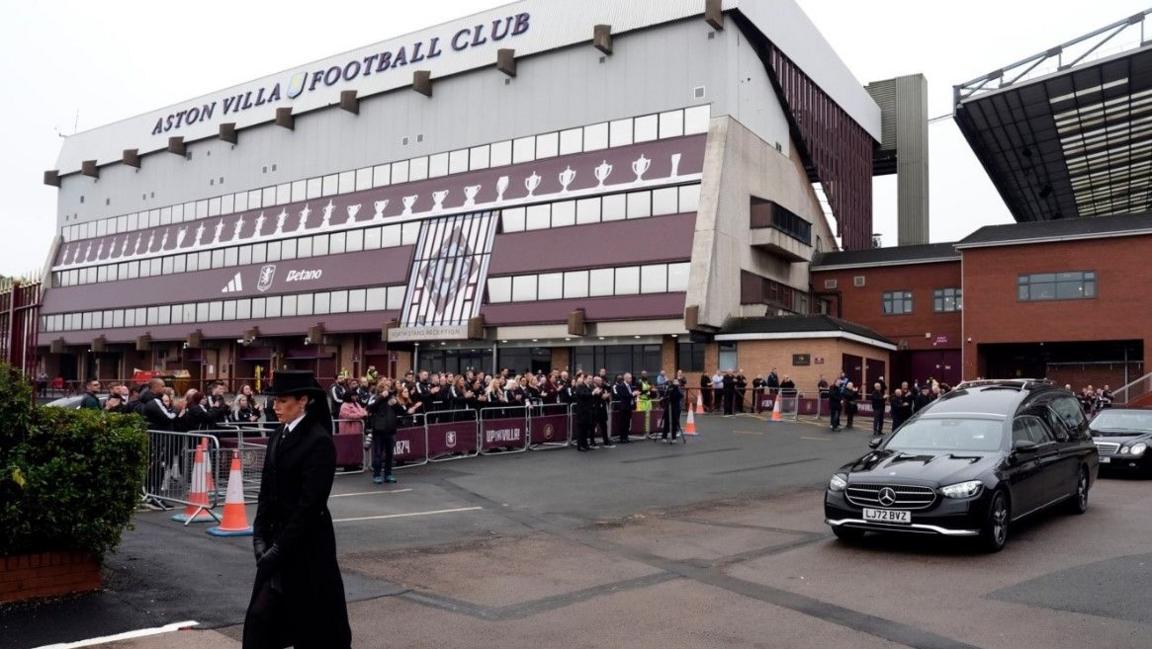 A funeral procession, led by a woman wearing a black hat with a veil down the back, makes its way past Villa Park with crowds gathered behind a barrier outside the stadium. The woman is followed by two black cars.