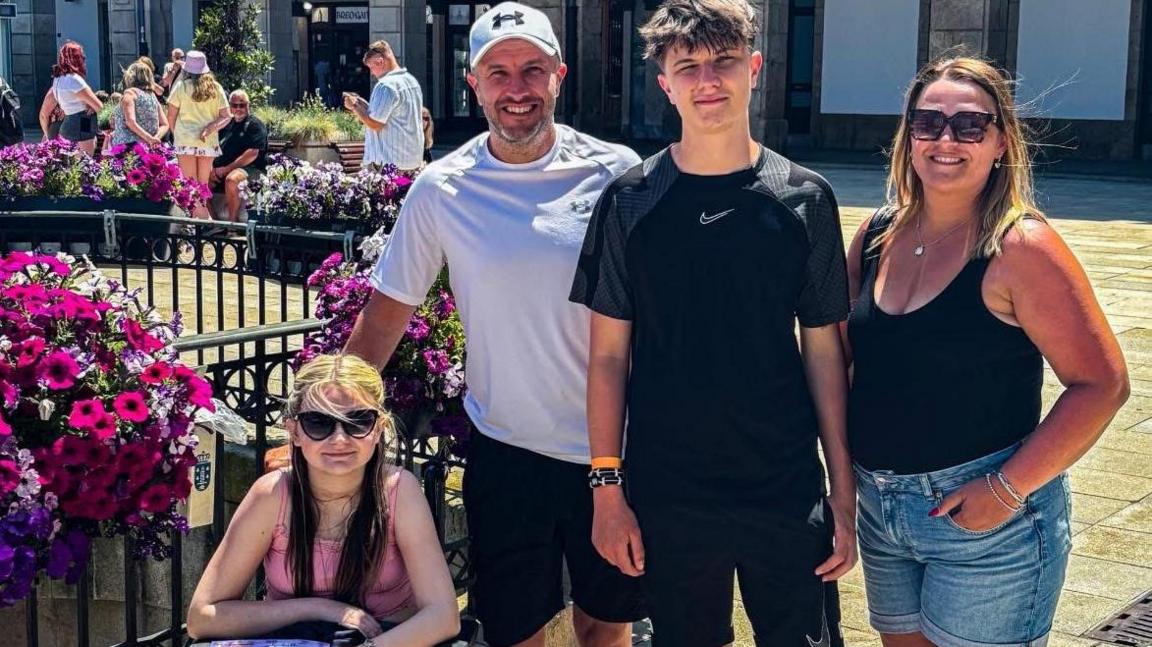 The family smile at the camera, Isla is wearing a pink top and is in a wheelchair