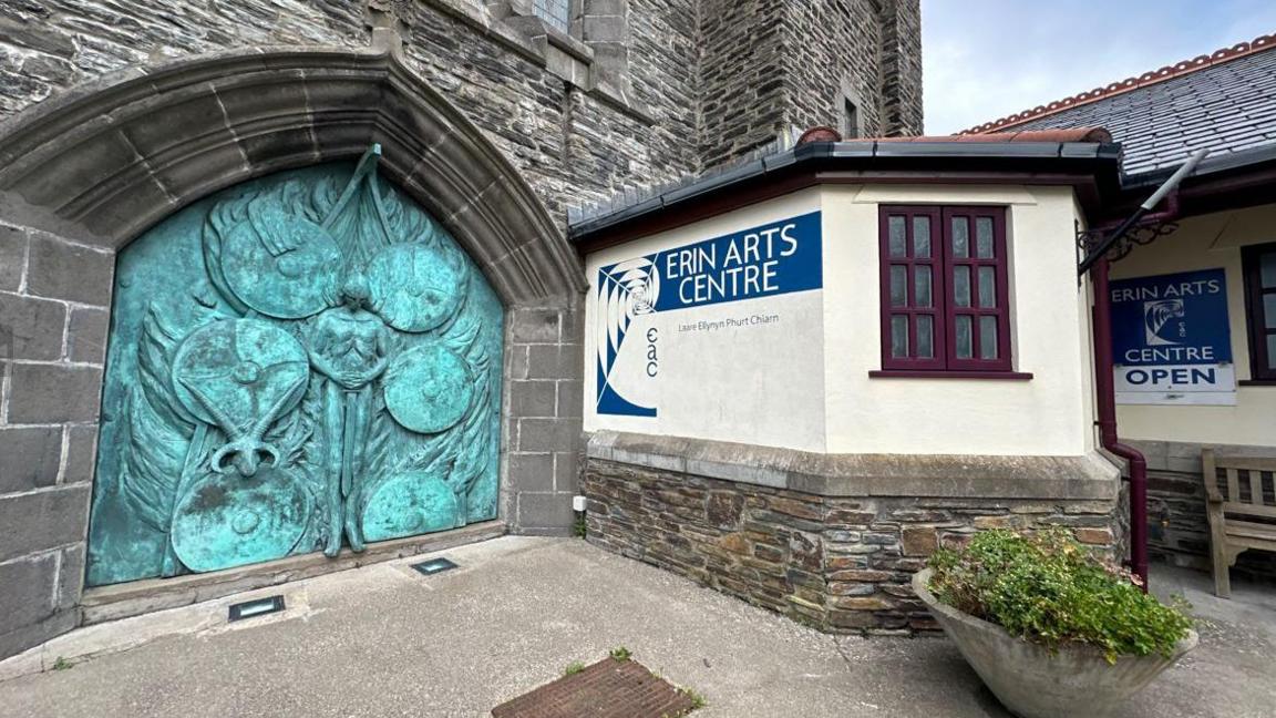 The entrance to the Erin Arts Centre. The name can be seen on blue mural on a cream wall. A piece of artwork shows a viking in a longboat at sea with shields surrounding him.