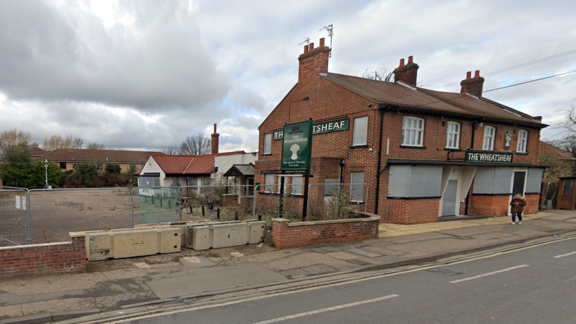 A photo taken from across the road facing the Wheatsheaf Pub, which has a green and white name board on the front and side of the building. There is an empty space to its left, with silver fencing around it. 