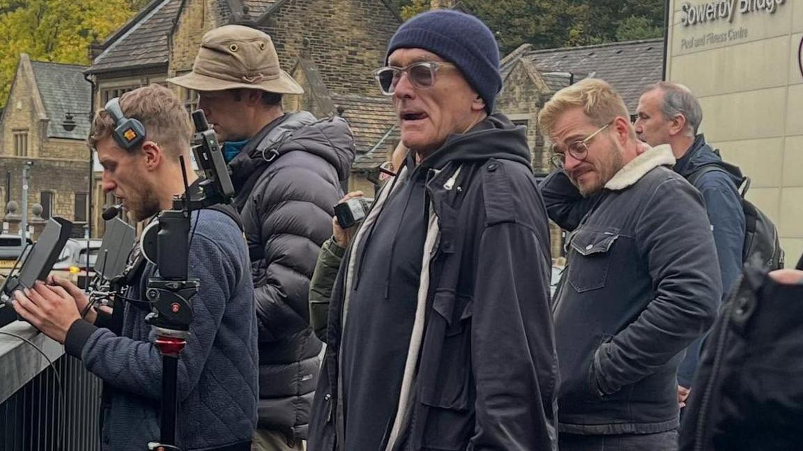 A group of five men dressed in navy blue coats, some wearing hats and some filming, as they stand on a bridge in Sowerby Bridge, West Yorkshire.