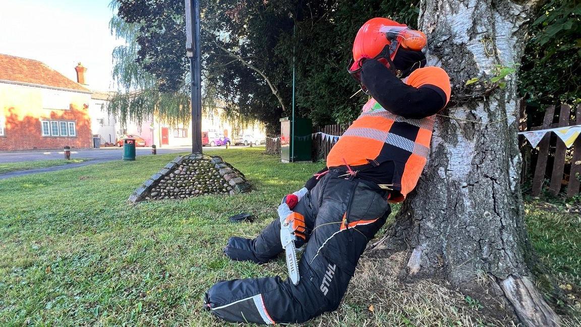 A scarecrow strapped to a tree. It is wearing an orange high-vis jacket, an orange hard hat and black trousers. The scarecrow is holding a plastic chain saw.