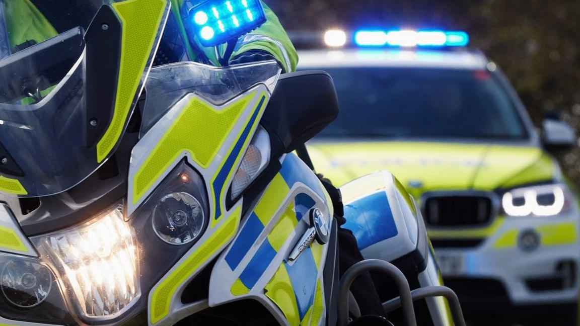 Close up image of a yellow and blue police motorbike, with a police car in the background. Both have blue lights lit up, and the motorcycle has a BMW emblem on the side.
