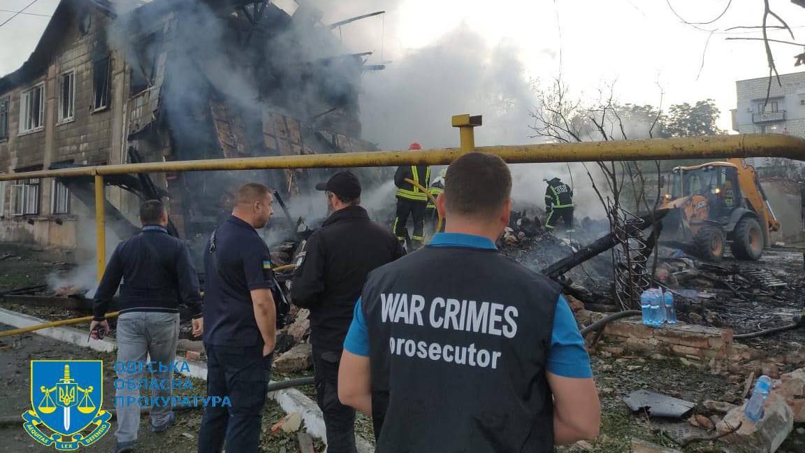 Odesa Regional Prosecutors stand at the site of a Russian drone attack on the port city of Izmail. The man closest to the camera has a vest reading "WAR CRIMES prosecutor". They are looking at a house that has been partly destroyed, there are lots of blackened and charred ruins, and it's still smoking heavily. In the background are firefighters hosing down the smoking ruins with water, and there is also a digger-type vehicle off to the side.