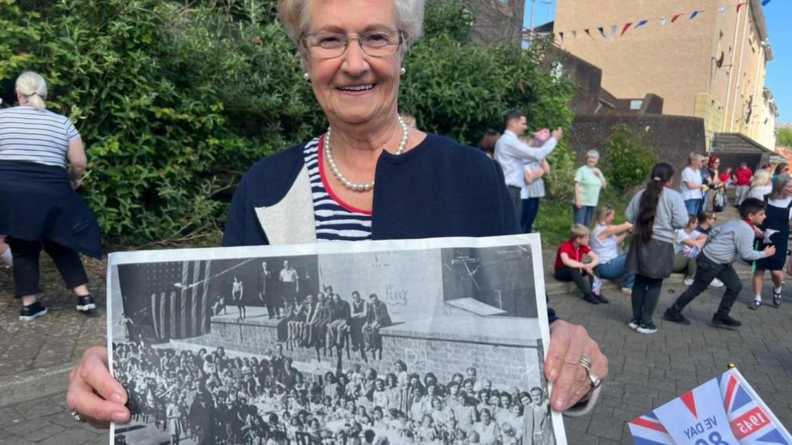 Helen Park is in a striped blue and white top and navy blazer.
She is wearing glasses and pearls.
She is holding a large black and white photograph.
A number of children are in the background.