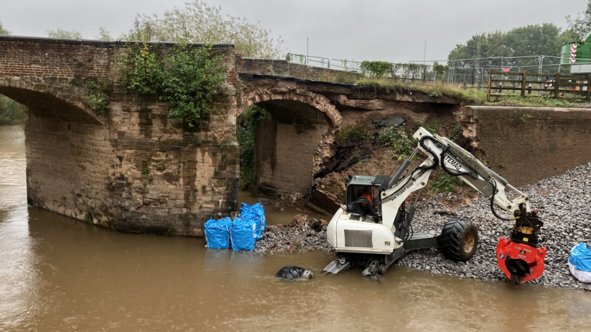 A small white crane is positioned beneath a small stone-and-brick bridge that has suffered damage to one side. 