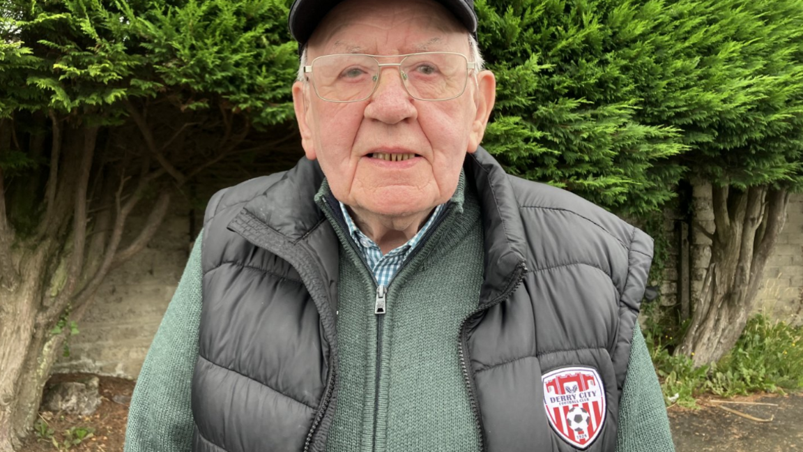 An elderly man looks into the camera. He is wearing glasses and a baseball cap with a green and white checked shirt, a light green zip-up cardigan, and dark sleeveless jacket with red-white-and-black Derry City logo on the breast. 