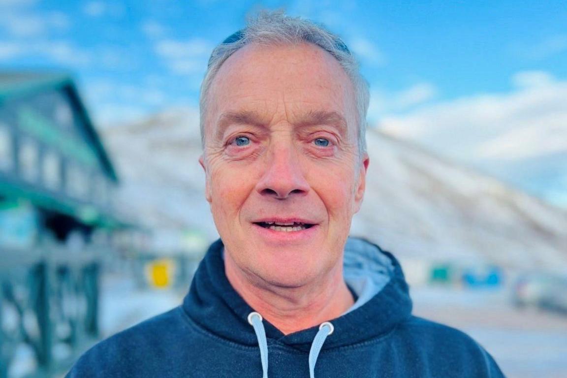 Man with grey hair, wearing a blue hooded top with the hood down, looking at camera, with a snowy scene behind and a cloudy blue sky.