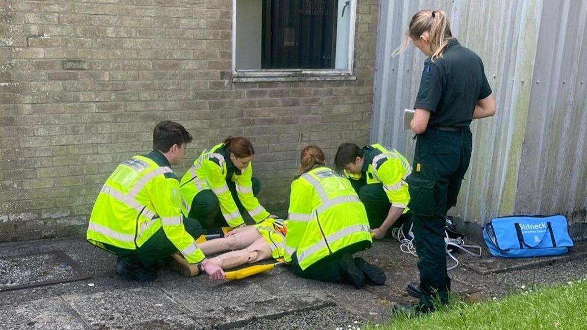 Myfyrwyr mewn cotiau llachar yn ymarfer delio gya chlaf