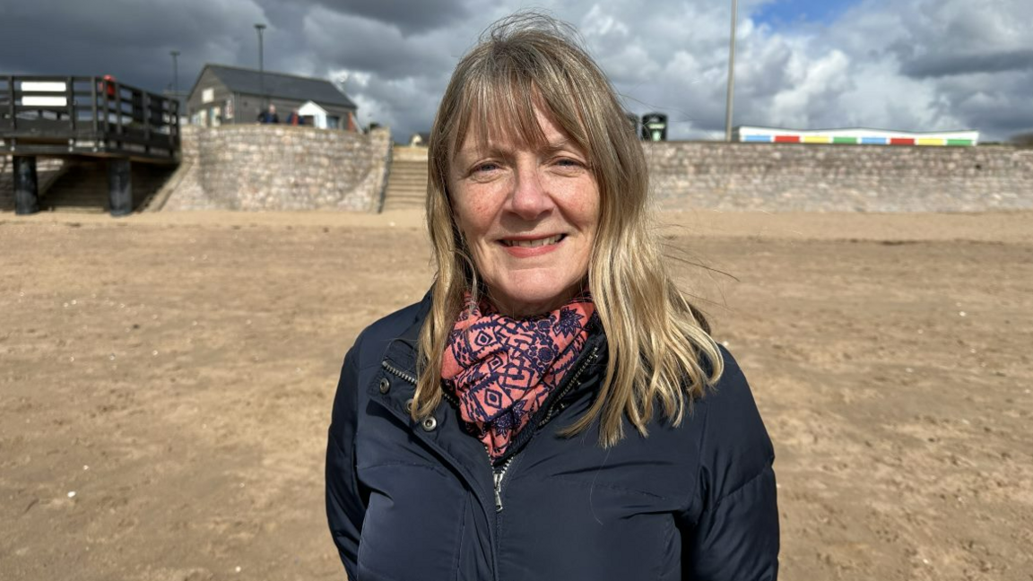 Marcia Fletcher is standing on a beach wearing a blue coat and a red and blue patterned scarf. 