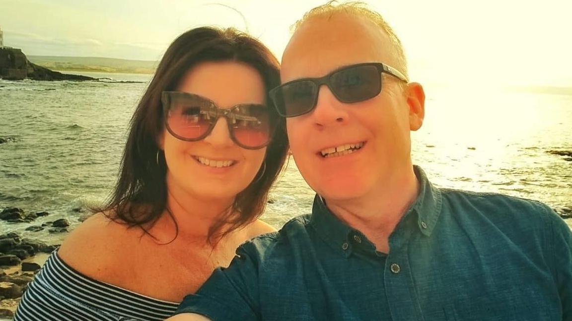 Larry and his wife pictured by the seaside both are wearing sunglasses and smiling into the camera with the sun shining behind them.