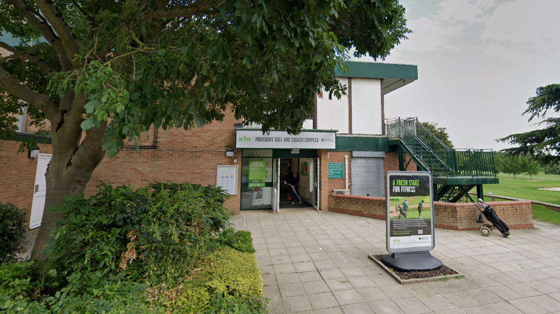 Entrance to a building, a golf course is visible in the background, there is a large green tree in the forground