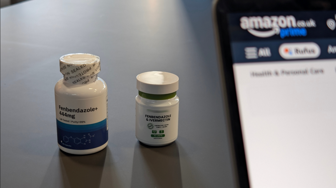 Two white pill bottles on a table, one labelled Fenbendazole and the other Ivermectin. A laptop stands beside them, with the Amazon logo visible on the screen.