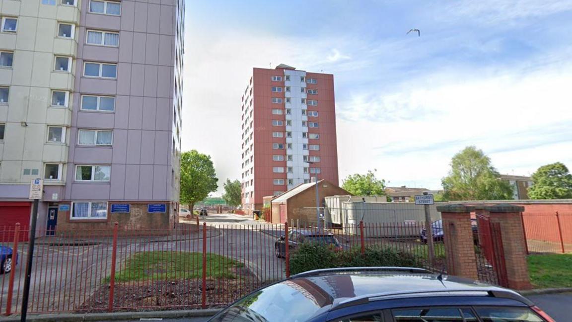 A Google Street view of two blocks of flat. The one of the left is purple and yellow and is cut off in the photo. The one on the right is red and white. It has multiple floors and windows on each floor. There are red railings and cars in view. 