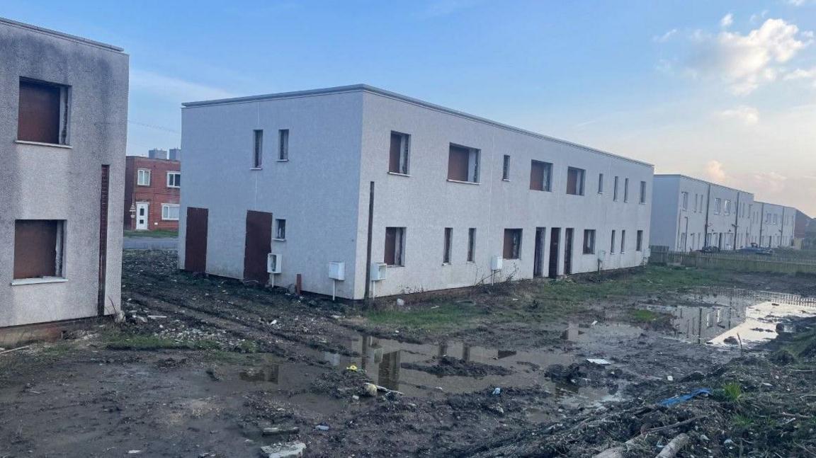 A series of grey flat-rooved block-buildings with small boarded out windows. The buildings are surrounded by muddy wasteland scattered with litter