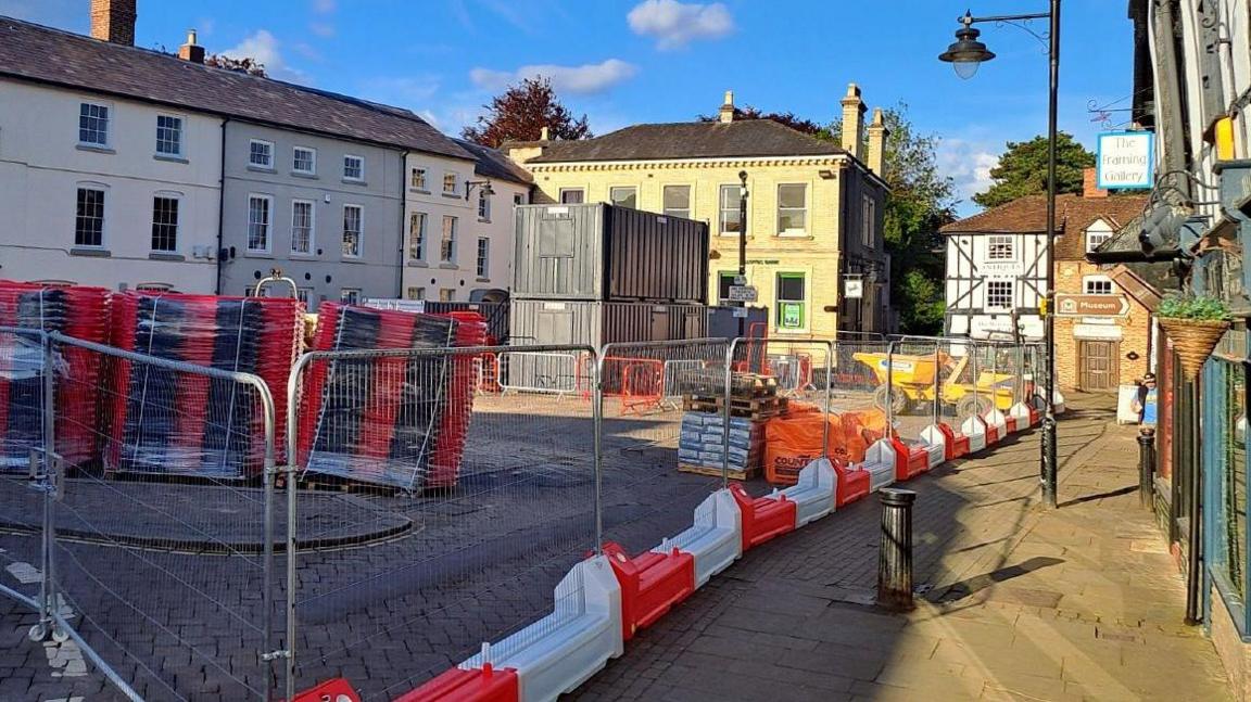 Red and white barriers, with fencing, contain construction materials in a half sunlit Leominster town square