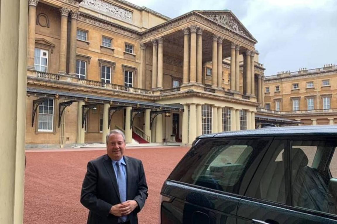 Jay Bloom, a middle-aged man with grey hair, wearing a navy suit and a blue shirt and tie, standing next to a dark coloured Range Rover in the quadrangle of Buckingham Palace, with its imposing portico behind him.