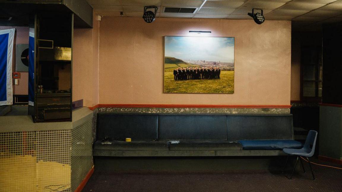 A room with a mirrored DJ booth on the left. There is some tatty seating at the back of the room and a photo of a group of men in dark clothing on the wall. To the right is a plastic chair