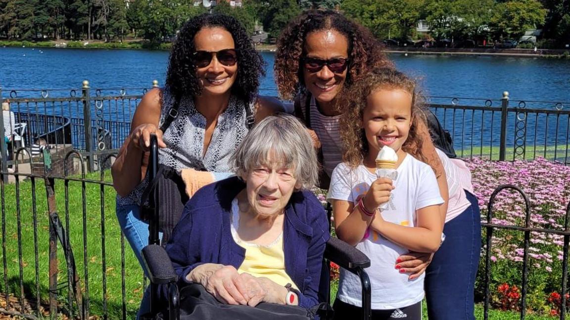 Joy Sesay, who has grey hair, is sat in a wheelchair wearing a yellow T-shirt and a blue cardigan. She is in the foreground surrounded by her family with a lake and a park full of flowers in the background. Behind Joy is a child wearing a white T-shirt and eating an ice cream on her left and two of her daughters, who black hair, jeans and colourful tops are behind her.