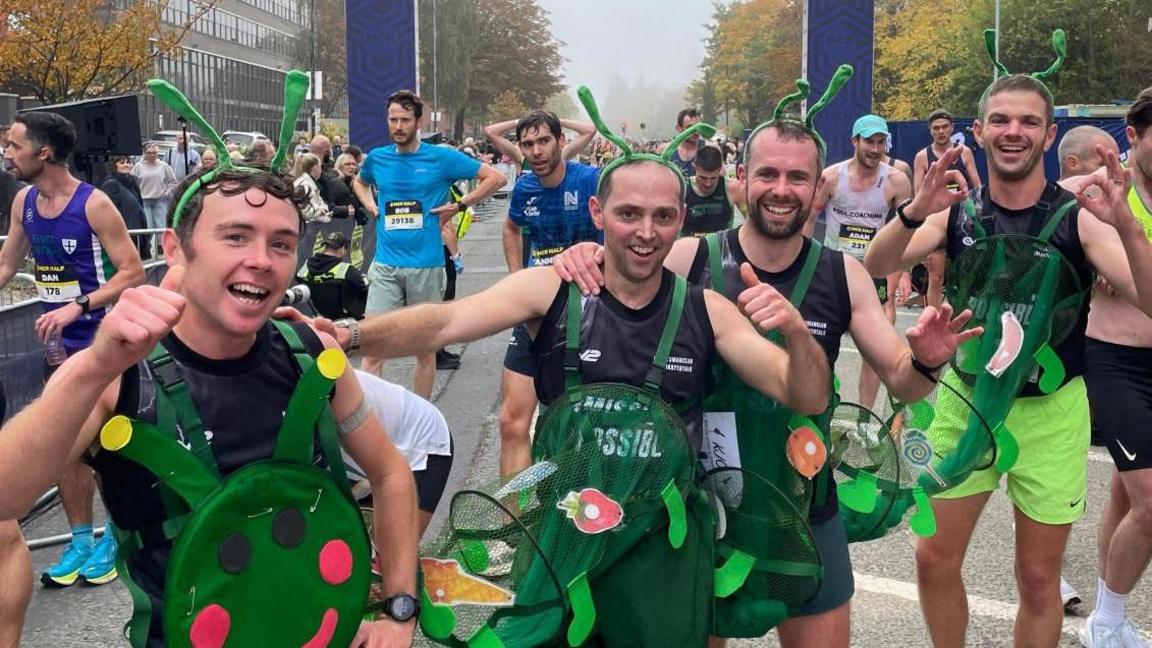 Group shot of four male friends smiling as they wear a green caterpillar costume and give thumbs up. They are at the finishing line with other runners coming behind them.