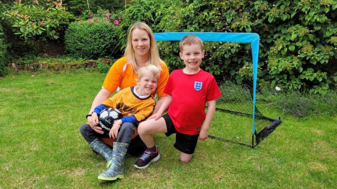 Emily Knight in a Holland football top kneeling down in front of a goal post in the garden with sons Edwyn, six and Wilfred, five holding a football