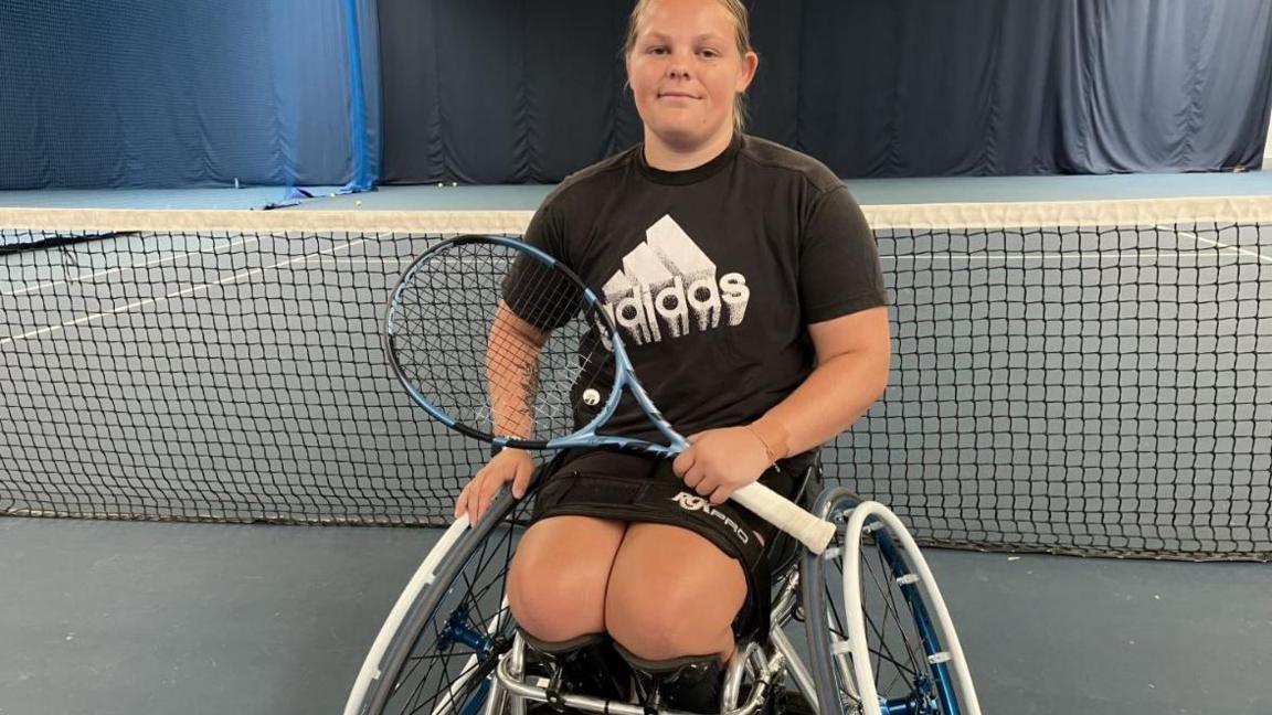 Ruby Bishop at a tennis court. She has blonde, swept-back hair and is wearing a black T-shirt with a large white Adidas logo. She is holding a tennis racket and looking directly at the camera. She is sitting in a specialist wheelchair.