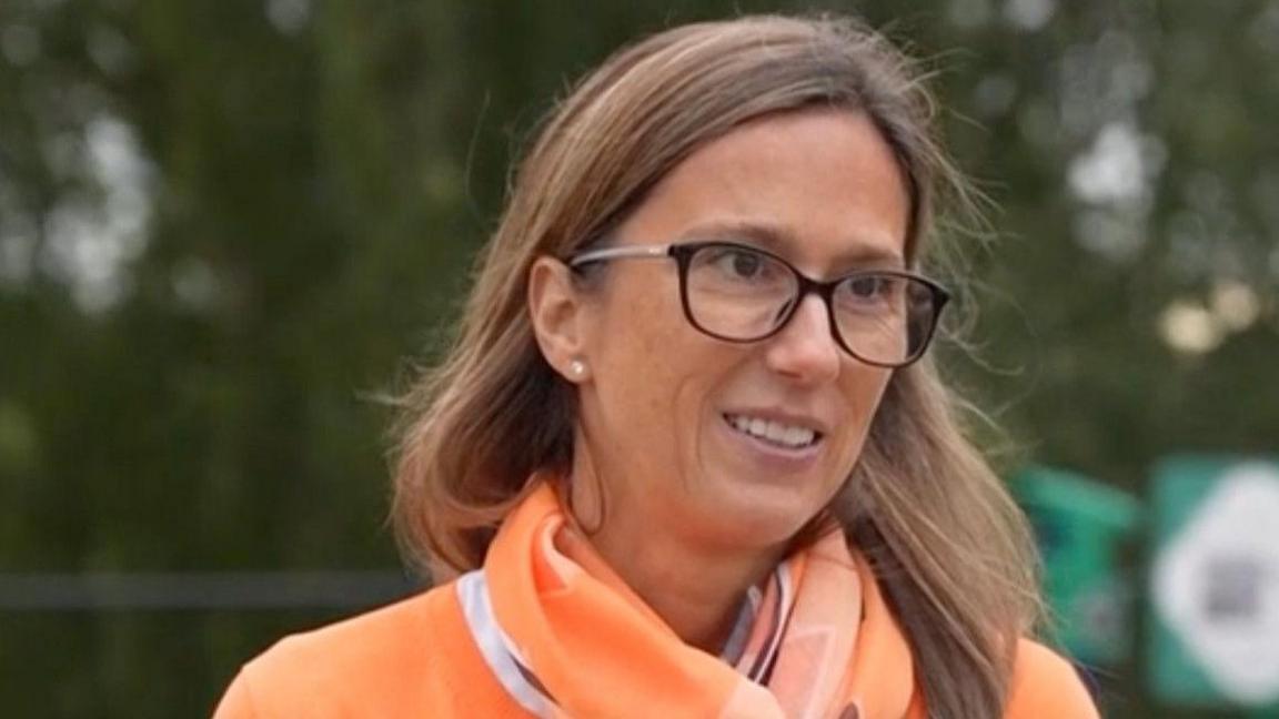 Kelly Richardson, who has long brown hair and is wearing glasses, wearing a bright orange coloured shirt and scarf in a park setting.