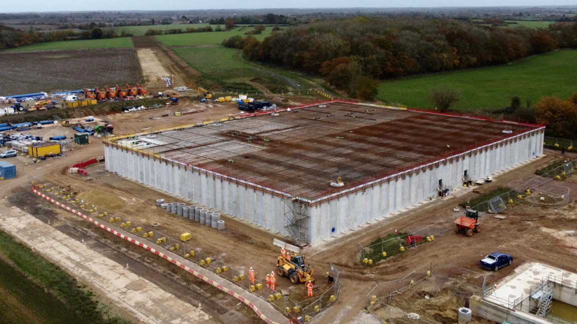 A drone image of the water storage reservoir and pumping station. A large square building is in the process of being built. Workers in orange hi-vis suits can be seen working around the site. Other building infrastructure can seen around the square building.