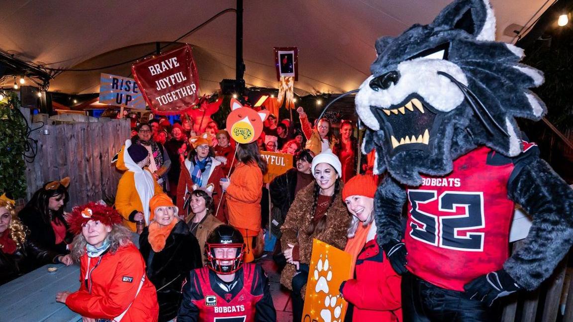 A large group of people dressed up in orange and holding up signs saying things like "brave, joyful, together" and "rise up". Some of the people are dressed up in American football attire and someone wearing the team mascot costume, a grey cat, stands at the side with their hands on their hips.