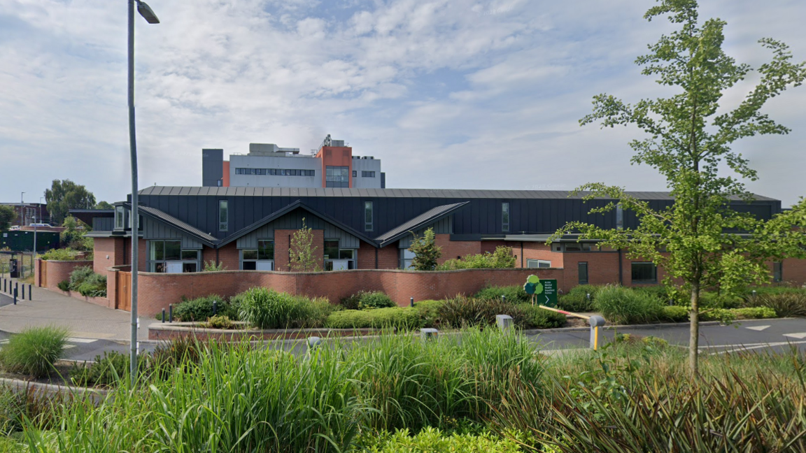 A single-story redbrick building with a black roof surrounded by bushes and trees and a road running outside it. In the background another building and above a blue sky with some clouds.