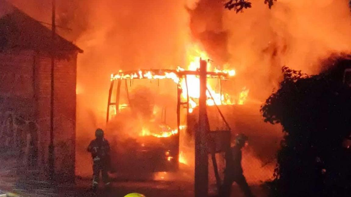 Flames surround the frame of what appears to be a bus, the entire scene is cast in an orange glow with a firefighter in the foreground.