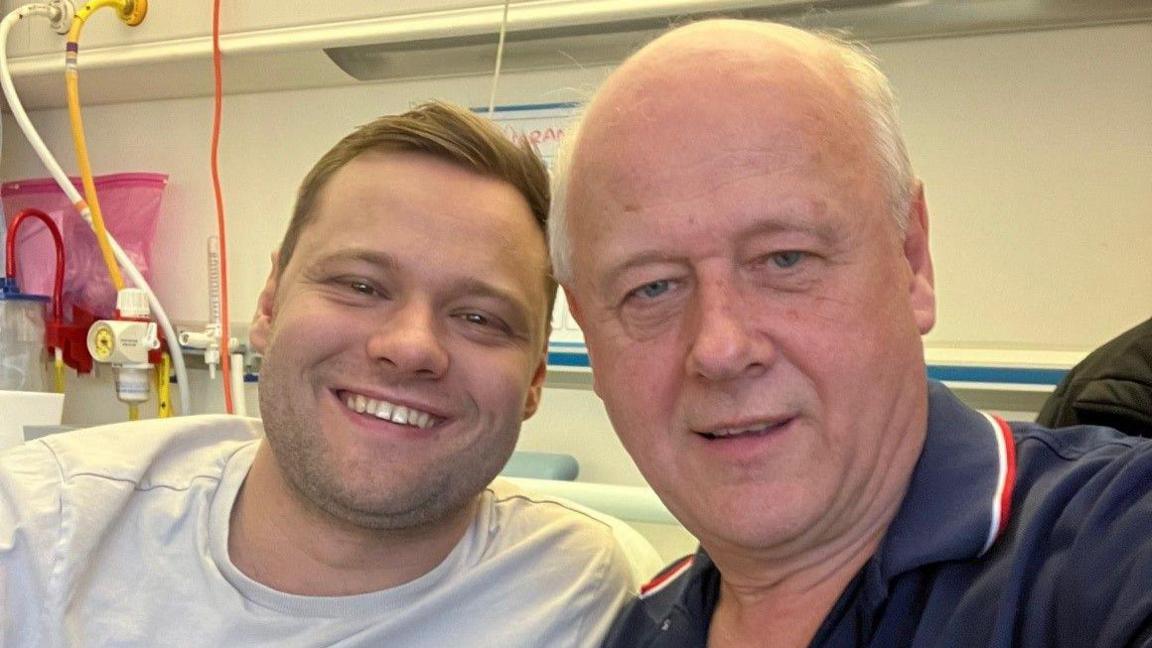 Kieran is smiling at the camera. He has short brown hair and is wearing a white t-shirt. Hi dad, Brian, is bald and is wearing a blue t-shirt.