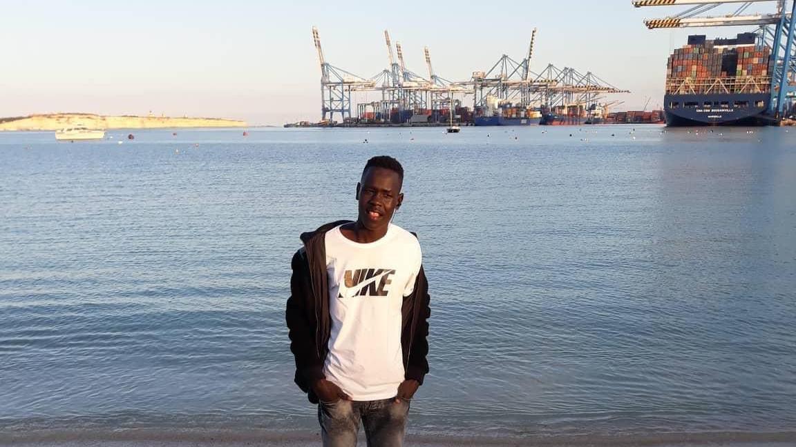 A young man in a white T-shirt, black jacket and jeans stands in front of a body of water with large ships and cranes in the distance.