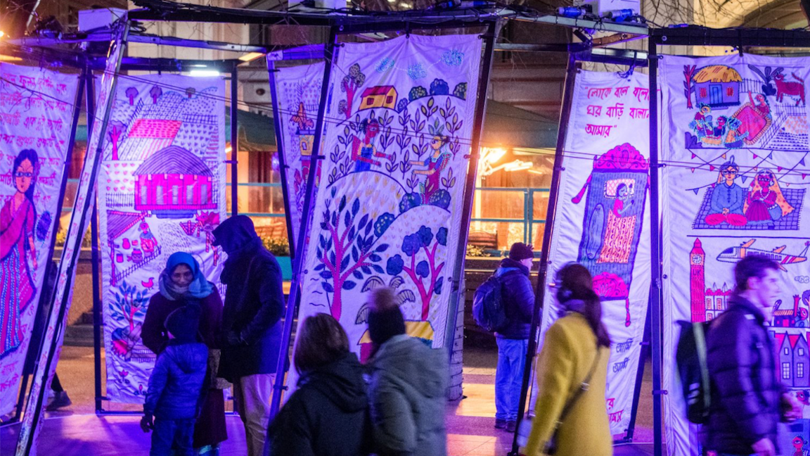 Numerous tapestries painted with vibrant purple colours, each depicting various different scenes. These scenes include houses, greenery and people. Numerous people can be seen walking in-between the tapestries.
