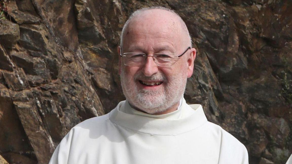 Fr John Murray. He is smiling at the camera. Fr Murray is bald and is wearing glasses. He has a white beard and is wearing white robes. Behind him is a rockface.