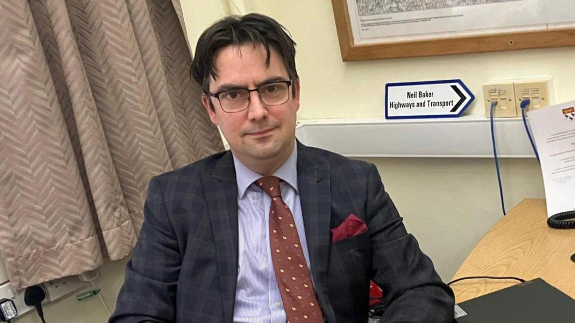 A man with dark hair wearing a navy suit and brown tie. He is sitting next to a sign which says "Neil Baker. Highways and Transport".