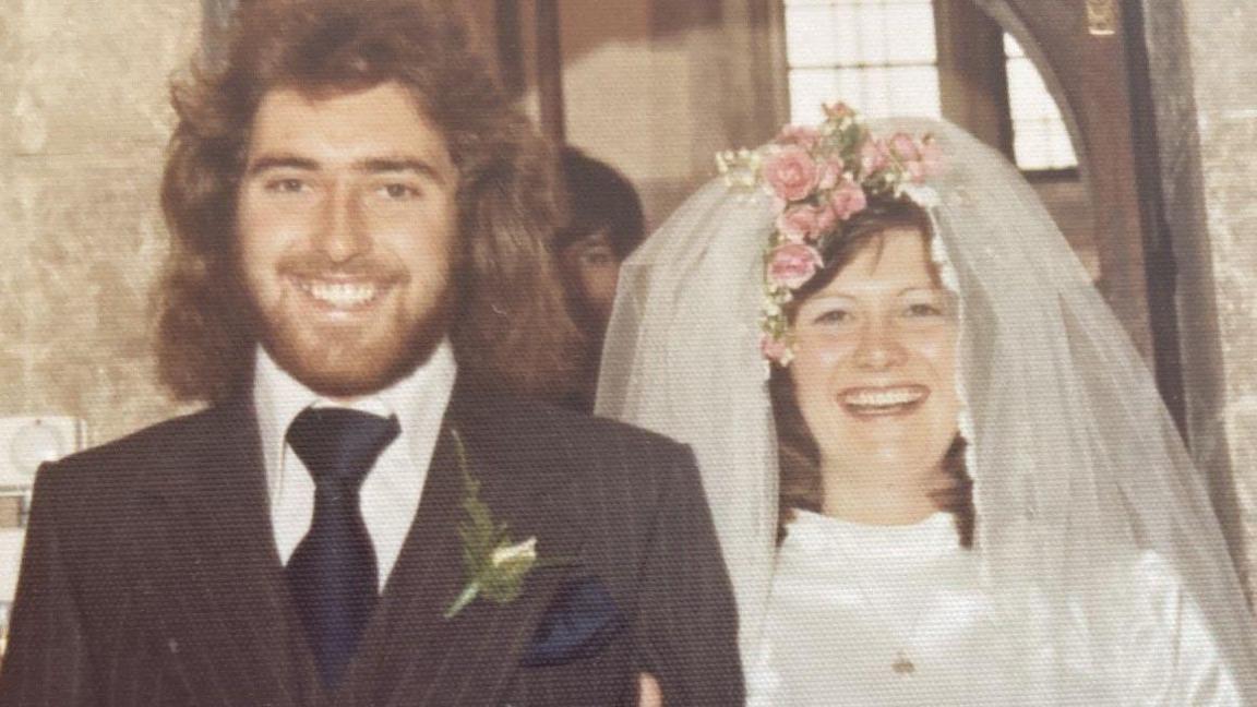 Wedding photo of Steve and Sandy Duckett in 1973. They smile at the camera in bridal outfits. Sandy wear a veil pushed back and held with flowers. 
